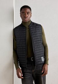 Zwarte gevoerde vest met een hoge kraag en rits, gedragen over een groene turtleneck. Kenmerkt een logo op de linkerzak en donkere jeans.