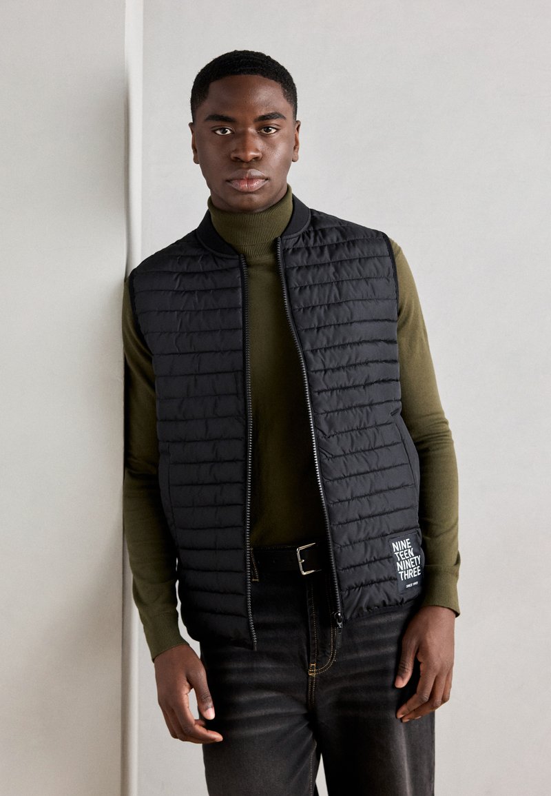 Zwarte gevoerde vest met een hoge kraag en rits, gedragen over een groene turtleneck. Kenmerkt een logo op de linkerzak en donkere jeans.