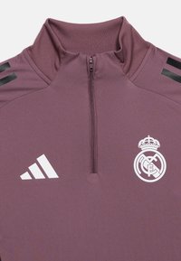 Μπορντό αθλητική μπλούζα με μισό φερμουάρ, κατασκευασμένη από λείο ύφασμα. Διαθέτει λευκό λογότυπο Adidas και έμβλημα της Real Madrid. Περιλαμβάνει τρεις μαύρες ρίγες στα μανίκια.