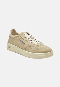 Sneakers in camoscio beige con dettagli traforati, chiusura con lacci e suola texturizzata. Presentano una toppa con logo e un design leggermente curvo.