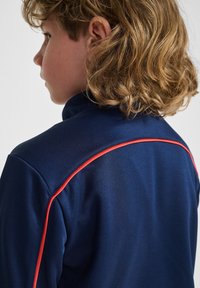 Marineblaue Trainingsjacke mit Stehkragen und kontrastierendem roten Besatz an den Schultern, gefertigt aus glattem, leichtem Material.