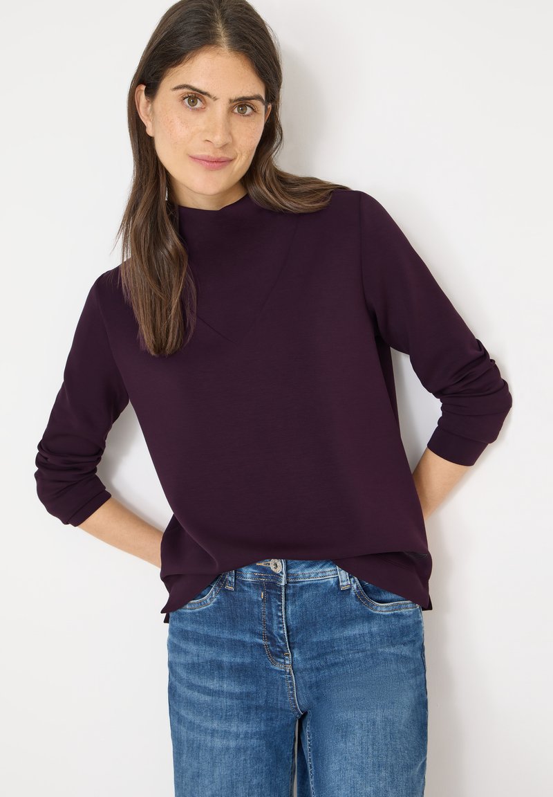 Violettes Langarmshirt mit hohem Ausschnitt, lockerem Sitz und lässigem Saum; kombiniert mit hellblauen Jeans.