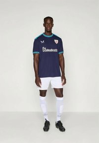 Ναυτικό ποδοσφαιρικό μπλουζάκι με ανοιχτό γαλάζιες λεπτομέρειες, που διαθέτει λογότυπο και κείμενο χορηγού, συνδυασμένο με λευκή βερμούδα και κάλτσες μέχρι το γόνατο. Μαύρα ποδοσφαιρικά παπούτσια.