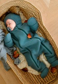 Miętowy polarowy kombinezon dziecięcy z zamkiem błyskawicznym, pluszowymi dodatkami na kapturze i ocieplanymi rękawiczkami, ułożony w wiklinowym koszu z jesiennymi liśćmi.