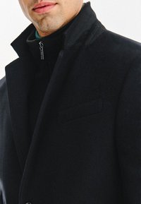 Czarny wełniany płaszcz z wysokim kołnierzem, o gładkiej teksturze, minimalistycznym designie i detalach w postaci bocznych kieszeni. Zamek błyskawiczny przy szyi.