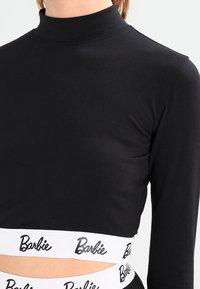 Svart långärmad crop top med hög hals och vit elastisk band med "Barbie"-logotyp. Slätt tyg, åtsittande design.