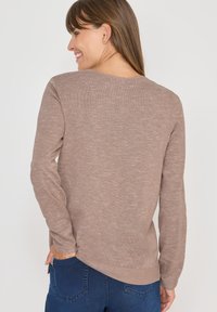 Maglione marrone a maniche lunghe in un tessuto morbido e texturizzato, caratterizzato da un colletto rotondo e spacco sui lati, abbinato a jeans blu scuro.
