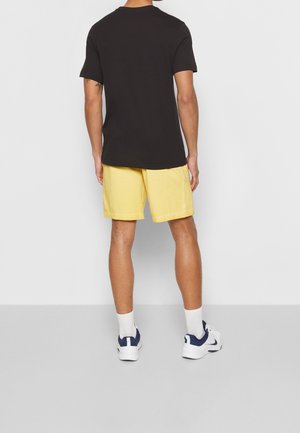 T-shirt en coton noir associé à un short jaune, avec une coupe décontractée. Des chaussettes blanches et des baskets bleu marine complètent la tenue.