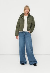 Olivovo zelená prešívaná bunda s cropped dizajnom, nosená cez krémový top a skombinovaná s modrými širokými džínami a čiernymi topánkami.