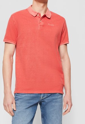 Homme portant un polo rouge délavé à deux boutons et un jean bleu clair sur un fond uni.