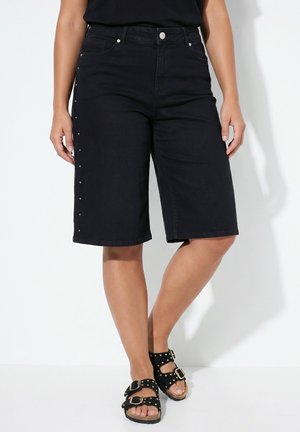 Personne portant un short en jean noir longueur genou avec des clous sur les côtés et des sandales noires à boucle, debout sur fond blanc.