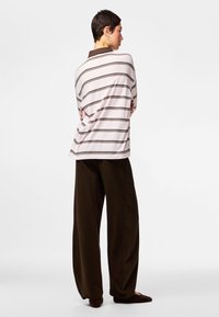 Osoba w luźnej, paskowanej koszuli z długim rękawem i szerokich ciemnych spodniach, stojąca z założonymi ramionami, zwrócona bokiem na jednolitym tle.