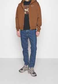 Brun zip-up hoodie över en svart grafisk t-shirt, blå jeans och grå sneakers med svarta accenter. Hoodien har framfickor och en logotyp.