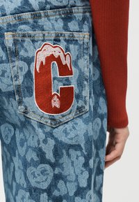 Πίσω τσέπη μπλε τζιν με σχέδιο και μεγάλο κόκκινο και λευκό κεντητό γράμμα "C", φορεμένα με εμφανές μανίκι σε σκουροκόκκινο χρώμα με ρίγες.