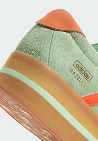 Zielone zamszowe sneakersy z pomarańczowymi akcentami, posiadające piętrową przezroczystą podeszwę oraz wytłoczone logo marki. Poziome paski w kontrastowych kolorach.