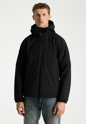 Junger Mann mit lockigem Haar, der eine schwarze Kapuzenjacke mit Reißverschluss und blaue Jeans trägt, steht vor einem einfarbigen hellen Hintergrund.