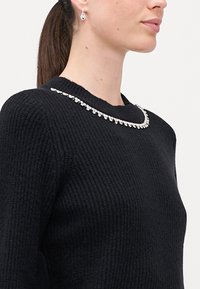 Μαύρο πλεκτό πουλόβερ με στρογγυλή λαιμόκοψη, διακοσμημένο με μια σειρά από διαφανείς ρουμπίνους κατά μήκος του γιακά. Ορατά ασημένια σκουλαρίκια σε σχήμα καρδιάς.