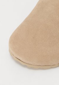 Chaussure en daim beige avec un bout rond et une surface lisse. La semelle en caoutchouc présente un bord festonné pour une meilleure adhérence.