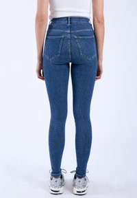 Höga blå jeans med smal passform, med två bakfickor och synlig sömnad, tillsammans med grå sneakers.