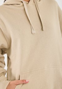 Beige hoodie med snore og en frontlomme, båret af en person med hånden hvilende i lommen.