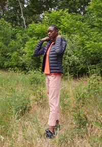 Zwarte gewatteerde pufferjack over een oranje top, roze cargobroek en zwarte schoenen, in een groene, grasrijke buitenomgeving.