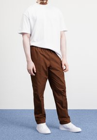 Person som bär en enkel vit t-shirt med kort ärm, bruna cargobyxor och vita sneakers som står på en blå heltäckningsmatta.