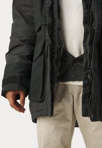 Parka-jakke i mørkegrøn, med lynlås foran, flere lommer og strukturerede manchetter. Indvendig foring synlig, som fremviser kontrastmateriale.