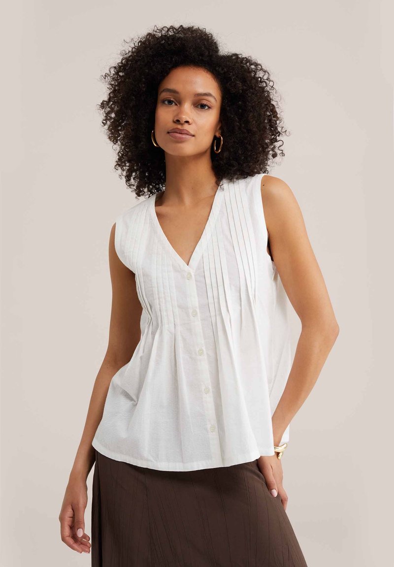 Woman with curly hair wears sleeveless white buttoned blouse with pleats, paired with a dark brown skirt, posing against plain background.
