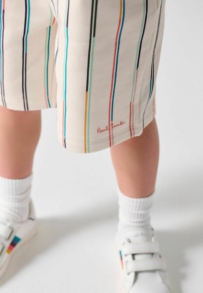 Barn iført beige shorts med flerfarvede lodrette striber, hvide sokker og hvide sneakers med farverige accenter.