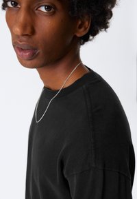 Musta puuvillapaita pyöreällä pääntiellä pehmeällä tekstuurilla, jossa on rento istuvuus ja tikatut yksityiskohdat. Kaulalla näkyy hopeaketjukaulakoru.