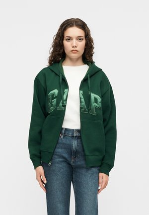 Femeie cu păr creț purtând un hanorac verde închis GAP peste un tricou alb și blugi albaștri, stând în fața unui fundal simplu deschis la culoare.