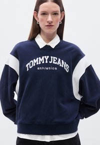 Μπλε σκούρο φούτερ με λευκές λεπτομέρειες, με κεντημένο το "TOMMY JEANS athletics" στο μπροστινό μέρος. Μακριά μανίκια και ριμπ τελείωμα.