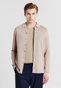 Beiger Cardigan mit kragen, Knopfverschluss und langen Ärmeln. Getragen über einem hellbraunen strukturierten Hemd, kombiniert mit dunklen Hosen.