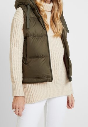Kvinde iført en olivengrøn puffervest over en cremefarvet ribstrikket sweater og hvide bukser mod en ensfarvet baggrund.