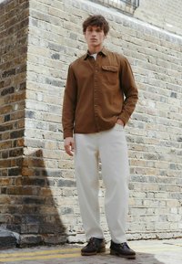 Hnedá košeľa na gombíky s vreckom na hrudníku, krémové nohavice a hnedé topánky s robustnými podrážkami. Postavený proti tehlovej stene.