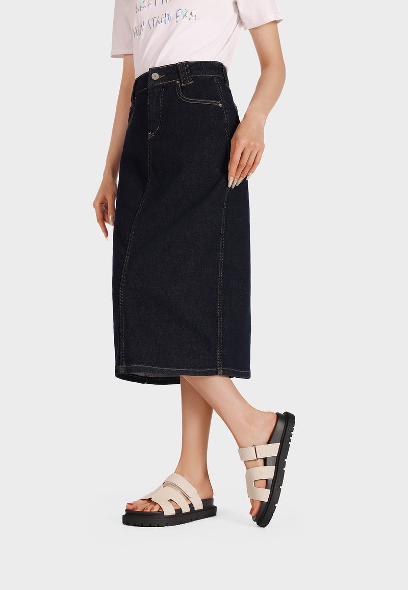 Gonna midi in denim blu scuro con cuciture a vista, abbinata a sandali beige con suola nera spessa e molteplici dettagli con fasce.
