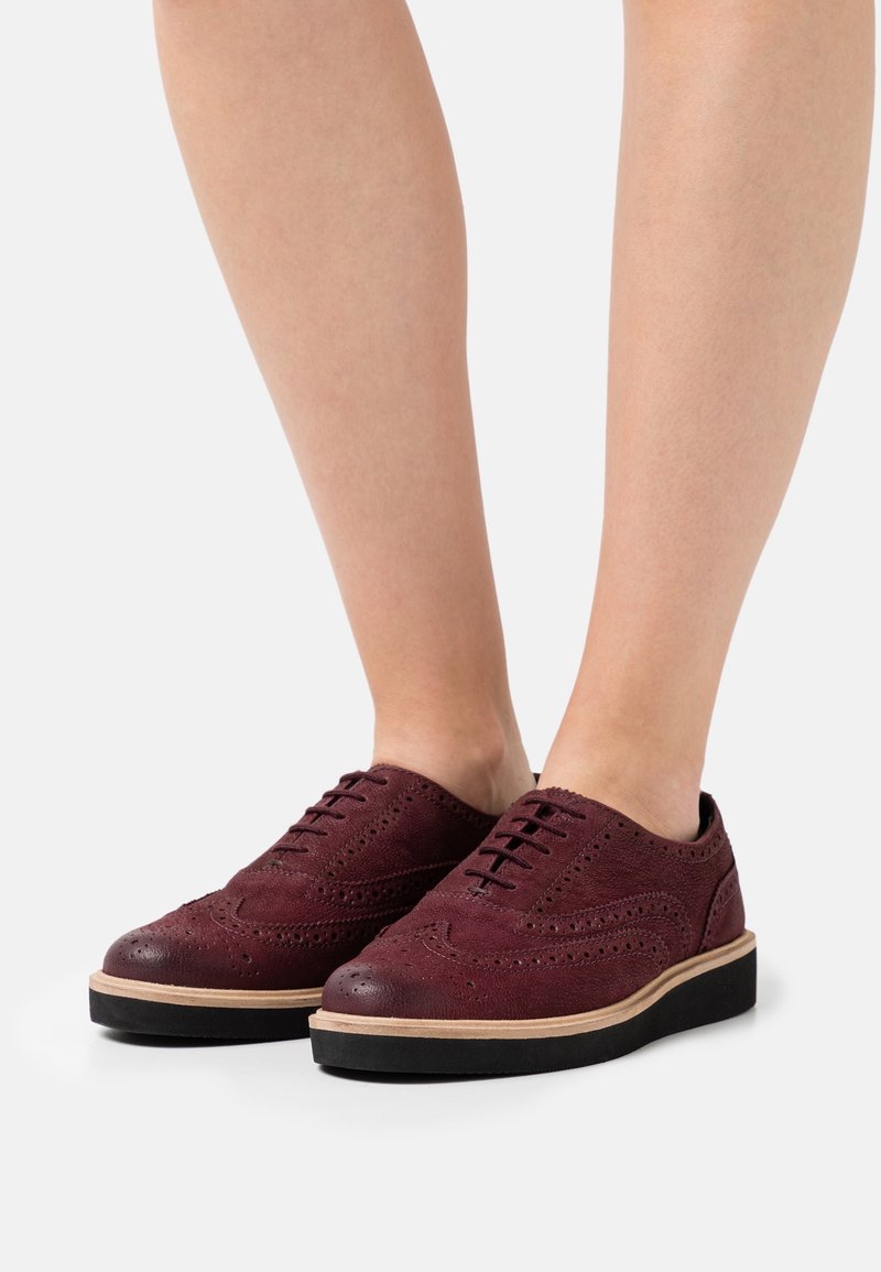 Jambes masculine portant des chaussures brogue à bout golf en cuir bordeaux avec semelles noires et bordure beige sur fond blanc.
