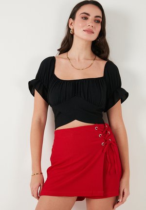 Zwarte crop top met puff mouwen en een vierkante halslijn, gecombineerd met een rode minirok met vetersluiting en oogjes aan de zijkant.