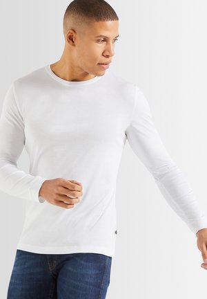 Weißes Langarmshirt aus glattem Baumwollstoff. Mit einem runden Halsausschnitt und taillierter Passform. Kombination mit blauen Jeans.