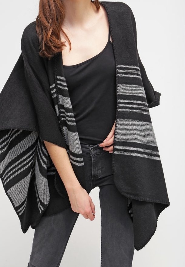 Femme portant un poncho en maille rayé noir et gris par-dessus un débardeur noir et un jean gris foncé, ajustant le poncho d'une main.