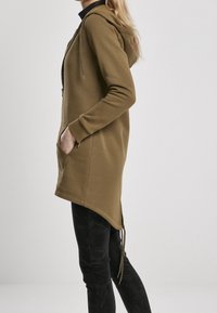 Olivově zelená mikina s kapucí, na zip, bočními kapsami a stahovací šňůrkou na spodním lemu. Má měkkou texturu a mírně prodloužený zadní díl.