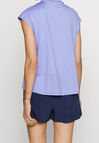 Personne portant un haut sans manches lavande et un short bleu foncé, vue de dos, debout devant un fond blanc uni.