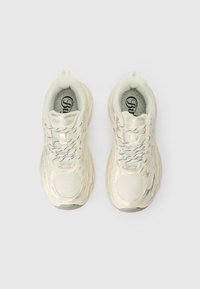 Fehér sportcipők, hálós felső résszel, texturált részletekkel és fényvisszaverő fűzőkkel. Vastag talp a párnázásért és stabilitásért.