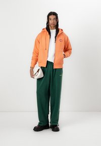 Orange hoodie with a front zipper and logo, white t-shirt, green sweatpants, and black chunky shoes. Holding a beige bag.