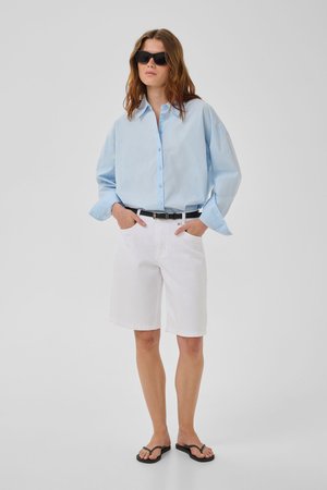 Žena nosiaca nadrozmernú svetlomodrú košeľu na gombíky, biele kraťasy po kolená s čiernym opaskom, čierne slnečné okuliare a čierne žabky.