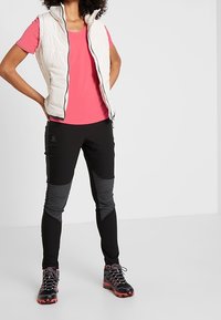 White padded vest with a zipper, bright pink short-sleeve shirt, black fitted pants with textured black panels, and black shoes with red accents.