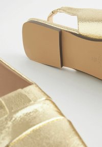 Złote buty z imitacji skóry o teksturowanym wykończeniu, z wyciętymi detalami i niskim słupkowym obcasem. Podeszwa ma neutralny kolor.
