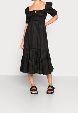 Femme portant une robe midi noire avec manches bouffantes et ourlet à volants, associée à des sandales noires, debout devant un fond clair uni.