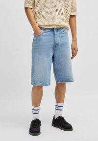 Pantaloni corti in denim blu chiaro con taglio dritto, cinque tasche e cuciture in vista. Indossati con scarpe da ginnastica nere e calze bianche con strisce blu.