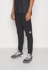 Melnas sporta bikses ar pieguļošu griezumu, ar elastīgu jostu un baltu Adidas logo kreisajā augšstilbā, apvienotas ar pelēkiem sporta apaviem.